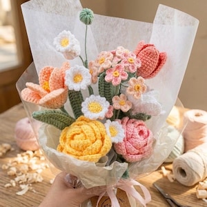 May include: A handmade crochet bouquet featuring a variety of colorful flowers. The bouquet includes yellow roses, pink tulips, white daisies, and other blossoms, all wrapped in paper and tied with a pink ribbon. The flowers are made of yarn.