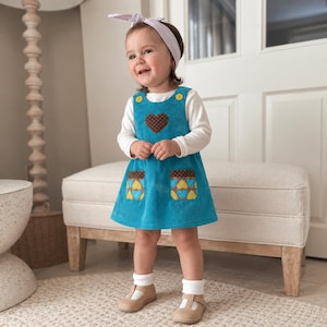 May include: A teal corduroy jumper dress featuring a brown heart applique with white dots on the chest, two front pockets with a brown and blue heart pattern, yellow buttons at the shoulders, worn over a long-sleeved white shirt with white socks.