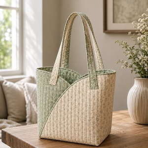 May include: A quilted tote bag featuring a diagonal colour-blocked design in sage green and cream, both adorned with a small-scale floral print. The bag has two matching fabric handles and vertical stitching throughout the quilted exterior fabric.