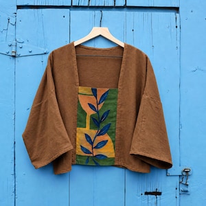 May include: A brown cropped jacket with wide sleeves and a square neckline. The center front panel features a hand-painted design of a dark blue leafy vine over a green and yellow abstract background, creating a distinct contrast.