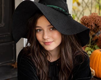 May include: A black witch hat featuring a wide, flexible brim and a pointed crown. The base of the crown is adorned with a dark green, textured ribbon trim that wraps around the hat, providing a subtle contrast to the solid black fabric.