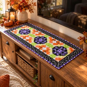 Könnte beinhalten: Ein rechteckiger, gesteppter Tischläufer mit einem sich wiederholenden Muster aus violetten, orangefarbenen und lindgrünen geometrischen Formen. Der Läufer ist mit einem schwarzen Stoff mit weißem Muster eingefasst und hat einen violetten Rand.