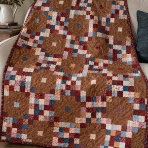 Puede incluir: Una colcha con un diseño de patchwork de pequeños cuadrados en tonos marrón, rojo oscuro, azul marino y rosa claro. El patrón consiste en bloques repetitivos de tela marrón sólida intercalados con secciones de cuadrícula multicolor.
