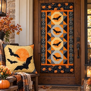 May include: A rectangular quilted wall hanging featuring a vertical pattern of four orange moons with black bat silhouettes, bordered by black fabric with orange pumpkins and skulls. A matching square throw pillow displays three black bats.