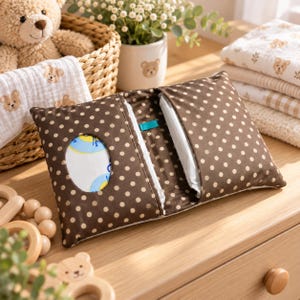 Peut inclure: Une pochette rectangulaire en tissu avec un fond marron et un motif à pois blancs. La pochette présente une ouverture ovale centrale pour distribuer des lingettes et une fermeture à rabat latéral. Une petite étiquette turquoise est visible.