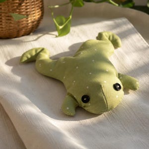 Può includere: Un peluche a forma di rana realizzato in tessuto verde chiaro con un motivo a pois bianchi. La rana presenta due occhi a bottone neri fissati saldamente sulla parte anteriore, con quattro arti divaricati e una testa arrotondata in tessuto.