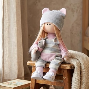 May include: A fabric doll with long blonde hair wearing a grey beanie with pink ear accents, a pink long-sleeved shirt, and grey and white striped overalls with a pink pocket. The doll holds a small grey plush toy and wears white sneakers.