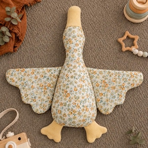 May include: A plush goose toy featuring a floral pattern with small yellow and blue flowers on a white background. The head, beak, and webbed feet are made of solid yellow fabric. The wings are extended outward, showcasing the detailed floral print.