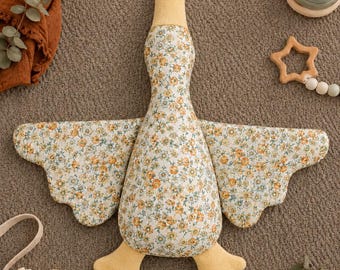 May include: A plush goose toy featuring a floral pattern with small yellow and blue flowers on a white background. The head, beak, and webbed feet are made of solid yellow fabric. The wings are extended outward, showcasing the detailed floral print.
