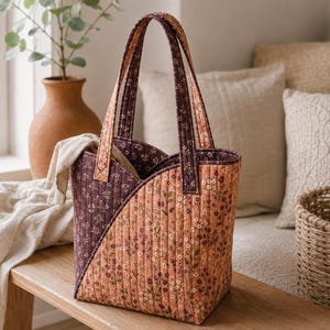 Op de afbeelding: Een gewatteerde tote bag met een diagonaal kleurblokontwerp. De ene kant is donkerpruim en de andere kant is gedempt perzik, beide versierd met een kleine bloemenprint. De tas heeft twee bijpassende stoffen handvatten en een verticaal stikpatroon.