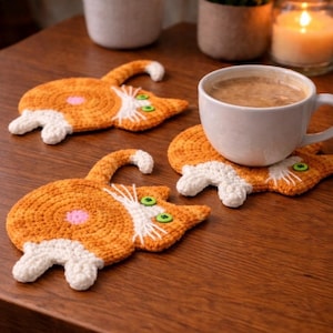 Peut inclure: Sous-verres faits à la main en crochet orange et blanc en forme de chat. Chaque dessous de verre présente un design de chat avec des yeux en boutons verts, une tache rose, une queue et des pattes blanches. Une tasse blanche avec du café est posée sur un des dessous de verre.