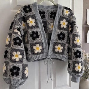 May include: A grey crochet cardigan with a floral design. The cardigan has a square pattern with black, white, and yellow flowers. It features long sleeves and a tie closure at the waist, offering a comfortable and fashionable style.