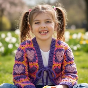 May include: A young girl smiles while wearing a handmade, colorful crochet cardigan. The cardigan features a heart pattern in shades of purple, pink, and yellow. The cardigan is tied with a purple bow. The girl is sitting outside on the grass.