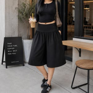 Peut inclure: Un haut henley noir à manches longues avec une encolure boutonnée, associé à une jupe-culotte noire taille haute dotée d'une large ceinture et de deux boutons latéraux. La tenue est complétée par des baskets noires et blanches et un sac à bandoulière marron.
