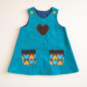 May include: A sleeveless turquoise corduroy pinafore dress featuring two yellow buttons at the shoulders. The front is decorated with a brown heart appliqué with blue polka dots and two pockets featuring a brown and blue heart pattern.