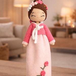 May include: A handmade crochet doll wearing a pink and white traditional Korean Hanbok dress. The doll has a brown bun with a floral crown and closed eyes. The dress features a white tie closure and embroidered flowers.