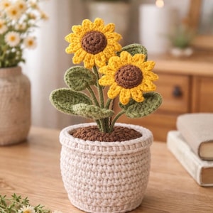 May include: A crocheted sunflower plant in a cream-colored pot. The sunflowers have yellow petals and brown centers, with green leaves. The pot is filled with brown material, resembling soil. The plant is on a wooden surface.