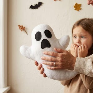 May include: A white plush ghost toy with a soft, rounded shape and two black oval eyes and a black oval mouth made of felt. The ghost has two outstretched arms and a curved bottom section, creating a simple, friendly, and recognisable Halloween-themed character.