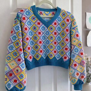 May include: A colorful, crocheted sweater with a V-neck design. The sweater features a repeating pattern of diamond-shaped squares in various colors, including blue, yellow, red, and pink. The sleeves and neckline are trimmed with a solid blue color.