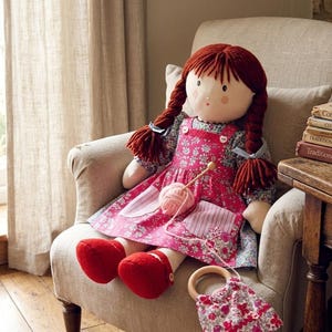 May include: A fabric doll with auburn yarn hair in two braids, wearing a floral print dress with a pink pinafore featuring two pockets. The doll holds a ball of pink yarn with a wooden needle and has red fabric shoes, sitting with a small floral bag.