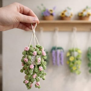 May include: A handmade crochet hanging planter with pink roses and green leaves. The planter is suspended by a cream-colored cord. Other crochet hanging planters with different flower and leaf designs are in the background.