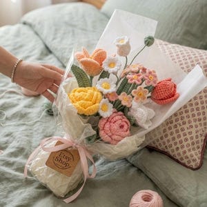 May include: A handmade crochet bouquet featuring yellow roses, orange tulips, white daisies, and pink flowers, wrapped in white paper and tied with a pink ribbon. A small tag with "Etsy Shop" is attached. The bouquet is presented on a bed with a green comforter.
