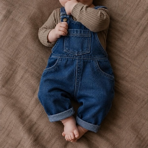 Può includere: Una salopette in denim blu scuro con tasca sul petto, tasche laterali e orli risvoltati, indossata sopra una maglia a maniche lunghe a coste color tortora. Il tessuto in denim presenta un lavaggio classico e bottoni metallici.