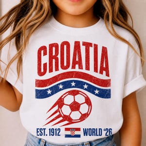 以下が含まれることがあります： 白い半袖Tシャツ。赤い文字で「CROATIA」と書かれ、その下に白い星が並ぶ青い波状の帯がある。さらにその下には、赤いサッカーボール、文字「EST. 1912」、クロアチアの国旗、「WORLD '26」がプリントされた、かすれた風合いのグラフィックデザイン。