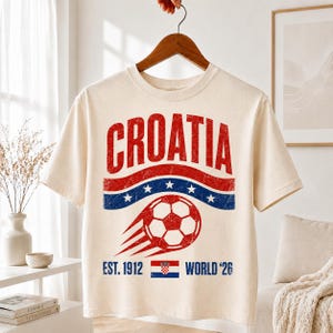 以下が含まれることがあります： クリーム色の半袖Tシャツで、赤、白、青のヴィンテージ風グラフィックがプリントされています。デザインには「CROATIA」の文字、動きのあるサッカーボール、白い星が描かれた青い帯、そして「EST. 1912 WORLD '26」というテキストが含まれています。