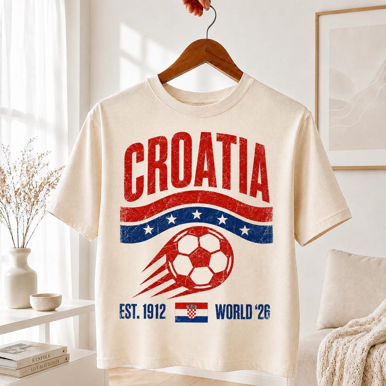 以下が含まれることがあります： クリーム色の半袖Tシャツで、赤、白、青のヴィンテージ風グラフィックがプリントされています。デザインには「CROATIA」の文字、動きのあるサッカーボール、白い星が描かれた青い帯、そして「EST. 1912 WORLD '26」というテキストが含まれています。