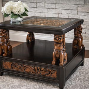 May include: Dark brown square coffee table with carved details and glass top. The table features carved lion-like figures as legs and a lower shelf with detailed relief carvings. A vase of white flowers sits on top.