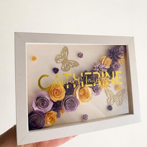 Peut inclure: Un cadre vitrine rectangulaire blanc contenant une composition de fleurs en papier dans des tons violets et jaunes. L'ensemble présente deux papillons dorés découpés et le texte doré CATHERINE et Merci Maîtresse.
