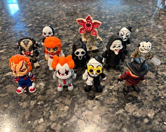 May include: A collection of eleven small horror-themed figurines including characters with orange hair, a black hooded figure with a white mask, a creature with a flower-like head, a figure in a striped sweater and hat, and a masked figure with a chainsaw.