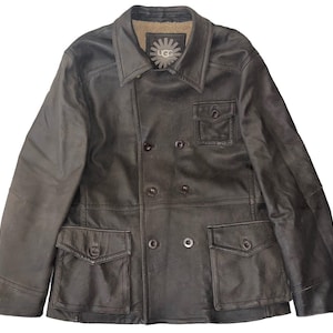 Puede incluir: Chaqueta de cuero gris oscuro con diseño de doble botonadura, con cuatro bolsillos delanteros y cuello con solapas. La chaqueta tiene un forro de borrego en el cuello y el logotipo de UGG. La chaqueta tiene una estética clásica y robusta.