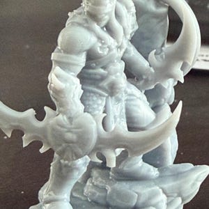 May include: A grey figurine of a warrior, holding two curved blades. The figure is standing on a rocky base, with detailed armour and a helmet. The design suggests a fantasy or gaming theme.