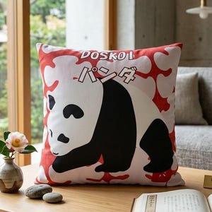 Puede incluir: Cojín cuadrado con una ilustración de panda. El panda es blanco y negro, sobre un fondo rojo y blanco. El cojín tiene el texto "DOSKOI" y caracteres japoneses. Está sobre una mesa de centro de madera.