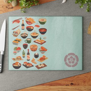 May include: A rectangular light blue glass cutting board featuring a variety of illustrated Japanese food items including sushi, ramen, and tempura on the left side, and a single mauve floral-shaped emblem on the bottom right corner.