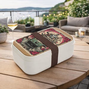 Puede incluir: Fiambrera rectangular blanca con tapa de madera que presenta una ilustración de estilo vintage de una mesa de billar, rosas y figuras. Una correa marrón asegura la tapa. La fiambrera está sobre una mesa de madera.