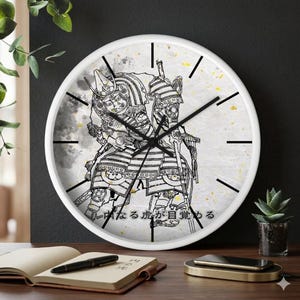 Op de afbeelding: Witte ronde wandklok met een zwart-wit illustratie van twee samoerai-krijgers. De wijzerplaat heeft zwarte uurmarkeringen en wijzers. Japanse tekst is zichtbaar aan de onderkant van de wijzerplaat.