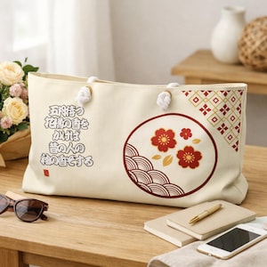 Puede incluir: Un bolso de tela color crema con asas de cuerda blanca. El bolso presenta un gráfico circular con flores de ciruelo rojas y doradas y patrones de ondas, un diseño geométrico en la esquina y texto en japonés impreso en color negro.