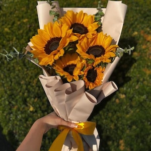 May include: A bouquet of sunflowers with bright yellow petals and dark brown centers, wrapped in beige paper and tied with a yellow ribbon. Green sprigs of eucalyptus are interspersed among the flowers. The bouquet is held against a blurred green background.