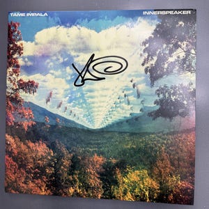 以下が含まれることがあります： Tame ImpalaのInnerspeakerの正方形のレコードジャケット。秋の木々の風景と、シュールな雲の形をした青い空が描かれています。中央には黒いインクのサインがあります。上部にはTame ImpalaとInnerspeakerの文字が白で印刷されています。
