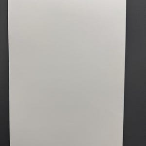 May include: A rectangular, off-white poster against a dark gray background. The poster has a smooth surface and a small, holographic sticker in the bottom right corner. The poster is blank, with no visible text or images.