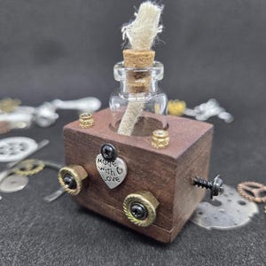 May include: A miniature steampunk-style oil lamp. A small glass bottle with a cork and wick sits atop a stained wooden block. The block has metal accents, including bolts and a heart-shaped charm that reads "Made with Love."