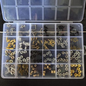May include: A clear plastic organizer filled with various small metal hardware pieces. The assortment includes screws, springs, eyelets, washers, and decorative elements in gold, silver, and black finishes. Ideal for crafting or small repairs.