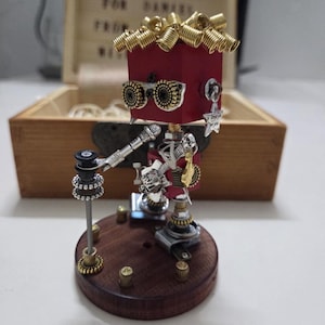 May include: A small robot figurine constructed from metal parts and a red cube head with gold coil hair. It features round metal glasses, a microphone on a stand, and a star-shaped charm. The figure stands on a circular wooden base with small metal accents.