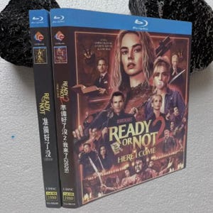 Könnte beinhalten: Eine Blu-ray-Hülle für den Film Ready or Not 2: Here I Come mit einer dunklen Collage aus mehreren Charakteren auf dem Cover. Der Titel ist in großen goldenen Buchstaben gedruckt, wobei der Text Ready or Not 2 Here I Come und chinesische Schriftzeichen zu sehen sind. Der Buchrücken zeigt den Titel und das Jahr 2026.