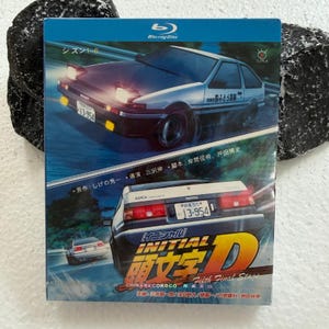 Op de afbeelding: Een Blu-ray hoes voor Initial D Fifth Final Stage met een witte en zwarte Toyota AE86 sportwagen in beweging op een weg. De hoes toont de auto vanuit voor- en achteraanzicht, Japanse tekst en de titel Initial D in grote oranje letters.