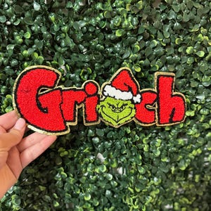 Op de afbeelding: Een rood en goudkleurig geborduurd embleem met het woord "Grinch". De "i" is vervangen door een groen Grinch-gezicht met een kerstmuts. De patch staat op een groene, bladerrijke achtergrond.
