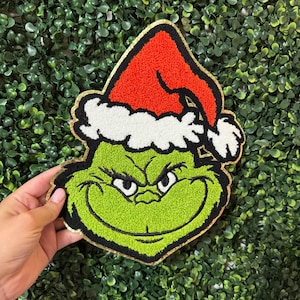 May include: A green Grinch face with a red Santa hat and white trim. The Grinch has a gold border and is held against a green leafy background. The Grinch's face has a mischievous expression.